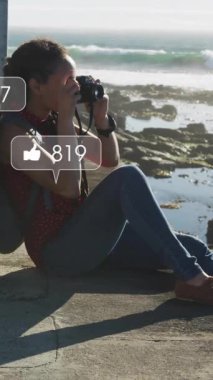 Okyanus kenarında oturup fotoğraf çeken bir kadın hakkında sosyal medya bilgilendirmeleri. Teknoloji, fotoğrafçılık, dijital, tatil, yaşam tarzı, seyahat