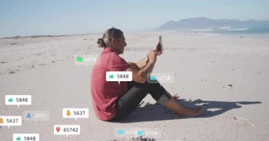 Kumsalda akıllı telefonları dinleyen, sosyal medya simgelerini etkinleştiren, artan nişan ölçümlerini belirten bir adam. Teknoloji, bağlantı, dijital, gevşeme, boş zaman, doğa, iletişim