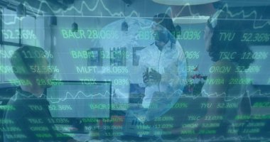 Presenting white male executive gesturing at meeting table, with monitors and stock-ticker overlay. Corporate, collaboration, digital, modern, professional, financial, teamwork