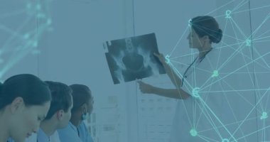 Doktor pelvis röntgeni yerleştiriyor ve stajyerler veri örtüsü kullanarak not alırken anatomiyi işaret ediyor. Tıp eğitimi, dijital sağlık, anatomi, eğitim, akıl hocalığı, klinik araştırma, takım çalışması