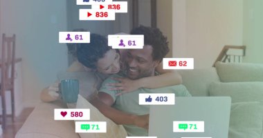 Kanepede kucaklaşan gülümseyen bir çift, dizüstü bilgisayarı kullanarak yüzen simgelerle geziniyorlar, ellerinde deniz mavisi kahve kupası tutuyorlar. Çift, samimiyet, rahatlama, dijital iletişim, rahat, modern, arkadaşlık