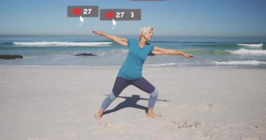 Sahilde savaşçı pozu veren kıdemli bir kadın sosyal medya ikonları kuruyor ve nişanı yükseltiyor. Dinginlik, farkındalık, sağlık, açık hava, denge, canlılık, motivasyon