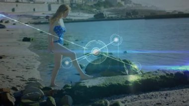 Kumlu kumsalda yosunlu kayaya basan bir kadın teknolojik demo için parıldayan ağ örtüsünü başlatıyor. Dijital, bağlantı, yenilik, fütüristik, doğadan esinlenilmiş, teknoloji, çevre
