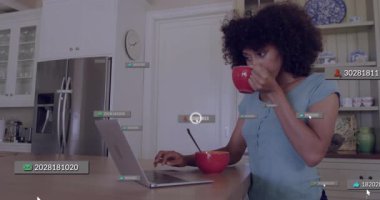 Tezgahta içki yudumlayan bir kadın dizüstü bilgisayarda yazarken yemek yiyor ve bitmeden önce teknolojik sinyal veriyor. Dijital uyarılar, uzaktan çalışma, modern mutfak, yaşam tarzı, teknoloji, çoklu görev, gündelik