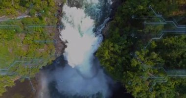 İnsansız hava aracı şelalenin tepesinde süzülüyor ve akıntılar boyunca iniyor. Devre tarzı HUD grafiklerini ortaya çıkarıyor. Kanyon, vahşi doğa, türbülans, dijital örtüşme, doğa, macera, keşif