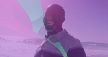 Kumlu sahilde polo tişört giyen, okyanus dalgaları ve mor kaplamalı gülümseyen olgun bir adam. Rahatlama, dinginlik, canlılık, rahatlık, açık hava, rahatlık, huzur.