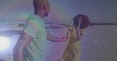 Yeşil tişört giyen ve kumsalda gülümseyen, okyanus dalgalı sarı elbiseler giyen çift el ele tutuşuyor. Romantizm, bağlantı, canlılık, dinginlik, macera, kaygısız, hayat dolu