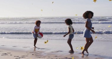 Anne ve iki çocuğu okyanus kıyısındaki kumlara altın konfetiyle plaj topu atıyorlar. Aile, eğlence, deniz kenarı, kutlama, eğlence, tatil, yaz