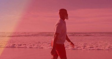 Adam kumlu sahilde köpüklü okyanus dalgalarıyla yürüyor, beyaz polo gömlek giyiyor, kırmızı atletik şort giyiyor. Plaj, yolculuk, sükunet, siluet, macera, manzara, rahatlık