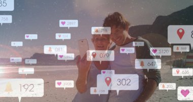 Anne-oğul ikilisinin gün batımında sahilde selfie çekmesi, akıllı telefon bildirim simgeleriyle. Aile, iletişim, rahatlık, açık hava, nostalji, sükunet, dijital