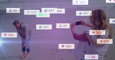 Ayak bileğindeki kumu fırçalamak ve akıllı telefon tutmak. Sosyal medya ikonlarıyla kumlu sahil şeridinde bir çift. Romantizm, bağlantı, açık hava, eğlence, teknoloji, huzur, yaşam tarzı