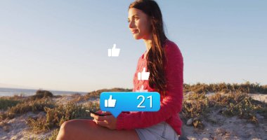 Kazaklı ve şortlu bir kadın kumulda akıllı telefon tutuyor. Başparmaklar yukarı, kopyalama alanı. Doğa, eğlence, teknoloji, sahil, açık hava, rahatlama, ruh hali.