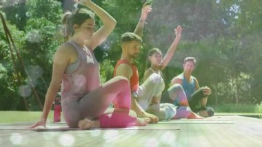 Eğitmen yoga grubunu minderlere yönlendiriyor. Gövdeleri omurga kıvrımları şeklinde döndürüp esnetiyor. Açık hava, sağIık, farkındalık, esneklik, doğa, sükunet, ahenk