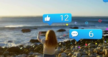 Gün batımında kayalık sahilde akıllı telefonu çerçeveletip sosyal medya ikonlarının canlı yayını güncellemesine neden olan bir kadın. Macera, etkili, açık hava, canlı, manzaralı, bağlantı, teknoloji