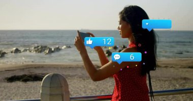 Akıllı telefon yetiştiren, deniz kenarını çerçeveleyen ve nişan büyümesini takip eden sosyal simgeleri canlandıran bir kadın. Deniz manzarası, teknoloji, yaşam tarzı, modern, iletişim, açık hava, bağlantı