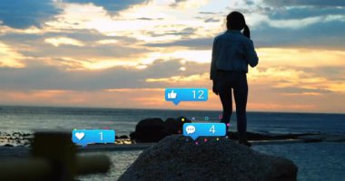 Gün batımında kayaya tırmanan kadın elinde telefonla sosyal medya ikonlarının yükselişini seyrediyor. Sahil, yalnızlık, doğa, macera, kutlama, dijital, sükunet