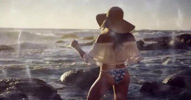 Gün batımında kayalık kıyı şeridinde yürüyen ve hasır şapkayı moda çekimi için ayarlayan manken. Deniz kenarı, gün batımı, doğal, boş zaman, zarafet, macera, siluet