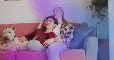 Kadın kırmızı koltukta uzaktan kumandaya basıyor ve partneri saçlarını okşarken ev eğlencesi başlatıyor. Samimiyet, rahatlık, arkadaşlık, rahatlama, yaşam tarzı, konfor, evcil