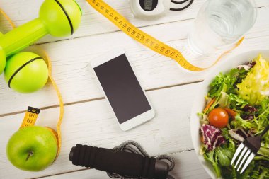 Overhead view of mobile phone and tape measure with dumbbells on wooden table