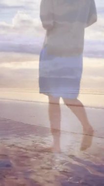 Sahilde gün batımında yalınayak yürüyen bir kadının arka görüntüsü.