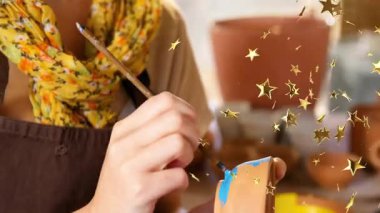 Bir kadın mavi boyaya daldırıyor ve çömleğin kenarındaki çömleği yıldız parlatıcısıyla süslüyor. El işi, yaratıcılık, zanaat, el işi, stüdyo, dekoratif, renkli