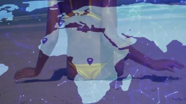 Sarı bikinili bir kadın kumsala yerleşiyor ve şeffaf teknoloji haritasında toplu iğne hatlarını gösteriyor. Plaj, sükunet, bağlantı, dijital, modern, açık hava, boş zaman