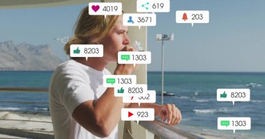 Plastik su şişesi ve sosyal medya ikonlarıyla metal korkuluklara yaslanan içki içen adam. Deniz manzarası, rahatlama, bağlantı, boş zaman, açık hava, minimalizm, refah