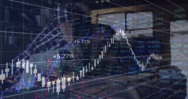 Analizci, ofis masasındaki monitör ve dizüstü bilgisayardaki pazar verilerini şeffaf çizelgelerle inceliyor. Finans, ticaret, analiz, veri görselleştirme, şirket, teknoloji, üretkenlik