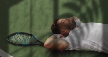 Tenis kortunda dinlenen tenisçi gömlek ve bileklikle uzanmış, yanında tenis raketi vardı. Spor, eğlence, açık hava, rahatlama, atletik, güneş ışığı, uyum.