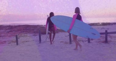 İki dişi sörfçü sörf yapmak için sörf tahtalarıyla çitlerin yanından geçiyor. Macera, arkadaşlık, sahil, deniz kenarı, dinlenme, sükunet, keşif