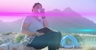 Bardağı yudumlayan ve tablet incelemesini başlatan bir kadın. Pembe mercek alevi gibi sıcak kamp alanında sürükleniyor. Yalnızlık, macera, açık hava, dinginlik, keşif, manzara, modern