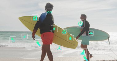 Sahilde sörf kıyafetleri içinde sarı ve yeşil sörf tahtaları taşıyan yürüyen yaşlı bir çift. Sahil, yaşlılar, eğlence, boş zaman, açık hava, rahatlama, kıyı