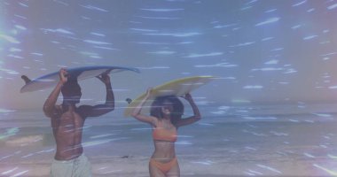 Kumsalda sörf tahtası taşıyan, dalgaları hisseden, şort ve turuncu bikini giyen yürüyen bir çift. Macera, rahatlama, deniz kenarı, boş zaman, canlı, enerjik, açık hava.