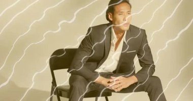 Moda reklamında değişen dalgalı ışıkları takip eden erkek manken sandalyeye yaslanıyor ve yakaları kavrıyor. Minimalist, zarif, sofistike, ruh hali, stüdyo, ışıklandırma, tefekkür