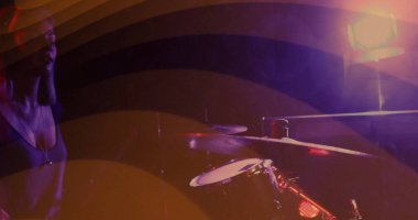 Mor-turuncu ışıklar altında sahnede davul seti ve ziller gösteriliyor ve müzisyen kolye takıyor. Performans, konser, ışıklandırma, atmosfer, müzik, sanatçı, sahne