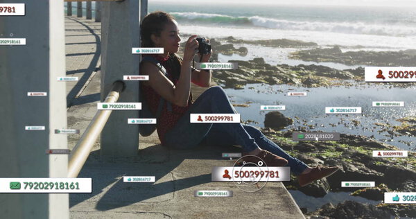 Сидящая латиноамериканка с камерой DSLR и обрамляющая береговую линию на прибрежной дорожке, с метками данных. Ландшафтный, приключенческий, фотографический, прибрежный, живописный, открытый, развлекательный
