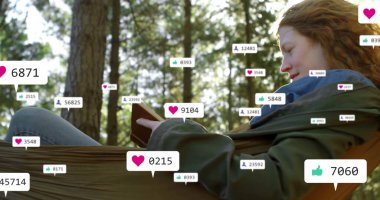 Güneşli çam ağaçları arasında, sosyal medya simgeleriyle yeşil hamakta kitap okuyan yaslanmış bir kadın. Doğa, rahatlık, rahatlama, açık hava, orman, dijital bağlılık, sükunet