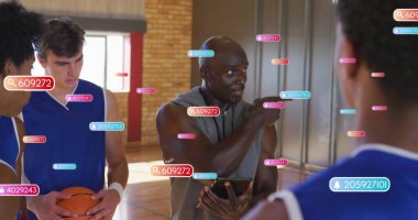 Kapüşonlu koç, elinde basketbol ve veri örtüleriyle sahadaki oyunculara el kol hareketi yapıyor. Atletik, takım çalışması, eğitim, performans, dijital, koçluk, spor