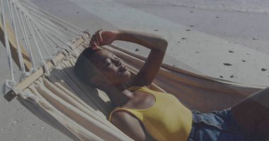 Sarı kolsuz bluz giyen, kumlu plaj hamağında gözleri güneşten koruyan, dalgalı bir kadın. Rahatlama, rahatlama, açık hava, yaz mevsimi, dinginlik, kıyı, yaşam tarzı