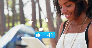 Orman kampında elinde tablet olan mavi çadırlı ve baş parmağıyla işaret eden bir kadın görülüyor. Kopya alanı. Doğa, macera, teknoloji, keşif, açık hava, boş zaman, bağlantı