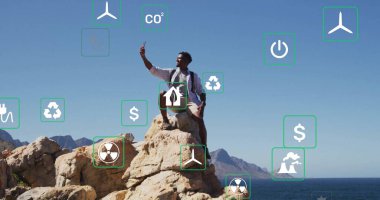 Diz çökmüş adam deniz kenarındaki kayalıklarda uçan yenilenebilir enerji ve tehlike simgeleriyle akıllı telefon kaldırıyor. Macera, keşif, sürdürülebilirlik, doğa, yenilik, çevrecilik, açık hava