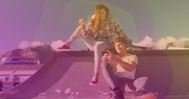 Su kenarındaki kaykay parkında ekose gömlekli oturan çift ellerinde akıllı telefon kaykay kulaklıkları tutuyor. Kentsel, yaşam tarzı, modern, boş zaman, açık hava, enerjik, canlı