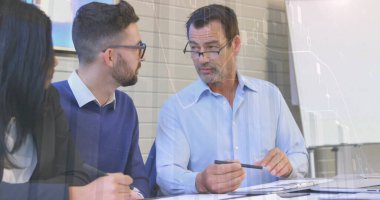 Mavi gömlekli kıdemli takım üyelerinden biri basılı belgelerle masa başında proje tartışması yapıyor. İşbirliği, takım çalışması, profesyonel, modern, ofis, strateji, beyin fırtınası