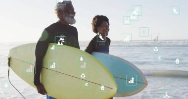 Dalgıç kıyafeti giyen ve sahilde sörf tahtası tutan açık mavi ve sarı renkli bir çift. Macera, boş zaman, kaygısız, açık hava, enerjik, canlı, uyum.