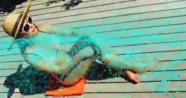 Güvertede turuncu havlunun üzerinde uzanmış uzanan, şapka kenarı parçacıkları titreşen bir kadın. Boş zaman, dinlenme, güneş banyosu, açık hava, yaz, sağlık, minimalizm.