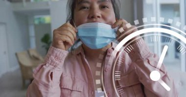 Olgun bir kadın ameliyat maskesi tutuyor ve oturma odasında saat grafiği izleme süresinde çıkartıyor. Yaşam tarzı, rahatlama, refah, ev, çağdaş, samimi, duygusal