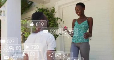 Adam sundurmada kırmızı ev anahtarı sunuyor, kadın alışverişi kutluyor, sosyal medya simgeleri yükseliyor. Kutlama, bağlantı, başarı, neşe, mutluluk, açık hava, modern