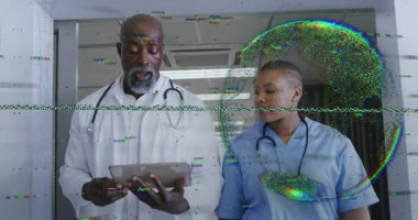 Doktor tableti tıklatarak hasta verilerini gösteriyor. Hemşireler koridoru takip edip bakımı tartışıyor. Tıbbi ekip, işbirliği, dijital teknoloji, sağlık, profesyonellik, klinik çevre