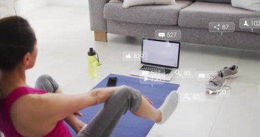 Kadın dizüstü bilgisayarda oynat tuşuna basıyor, sosyal simgelerle canlı bağlantı için minder üzerinde vücut çalışması yapıyor. Fitness, sağlık, ev, aktif teknoloji, motivasyon, disiplin