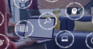 Meslektaşı kanepenin arkasından çıkarken bir yandan da dizüstü bilgisayarda eğitim ikonlarını süren bir kadın. İşbirliği, dijital arayüz, modern ofis, birlikte çalışma, teknoloji, dinamik, profesyonel